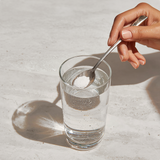 Hand adding collagen to a glass of water on a light surface