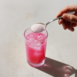 Hand adding pink powder to a glass of pink liquid with ice cubes on a light background