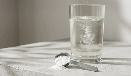Glass of water and a metal scoop of creatine powder on a kitchen counter.