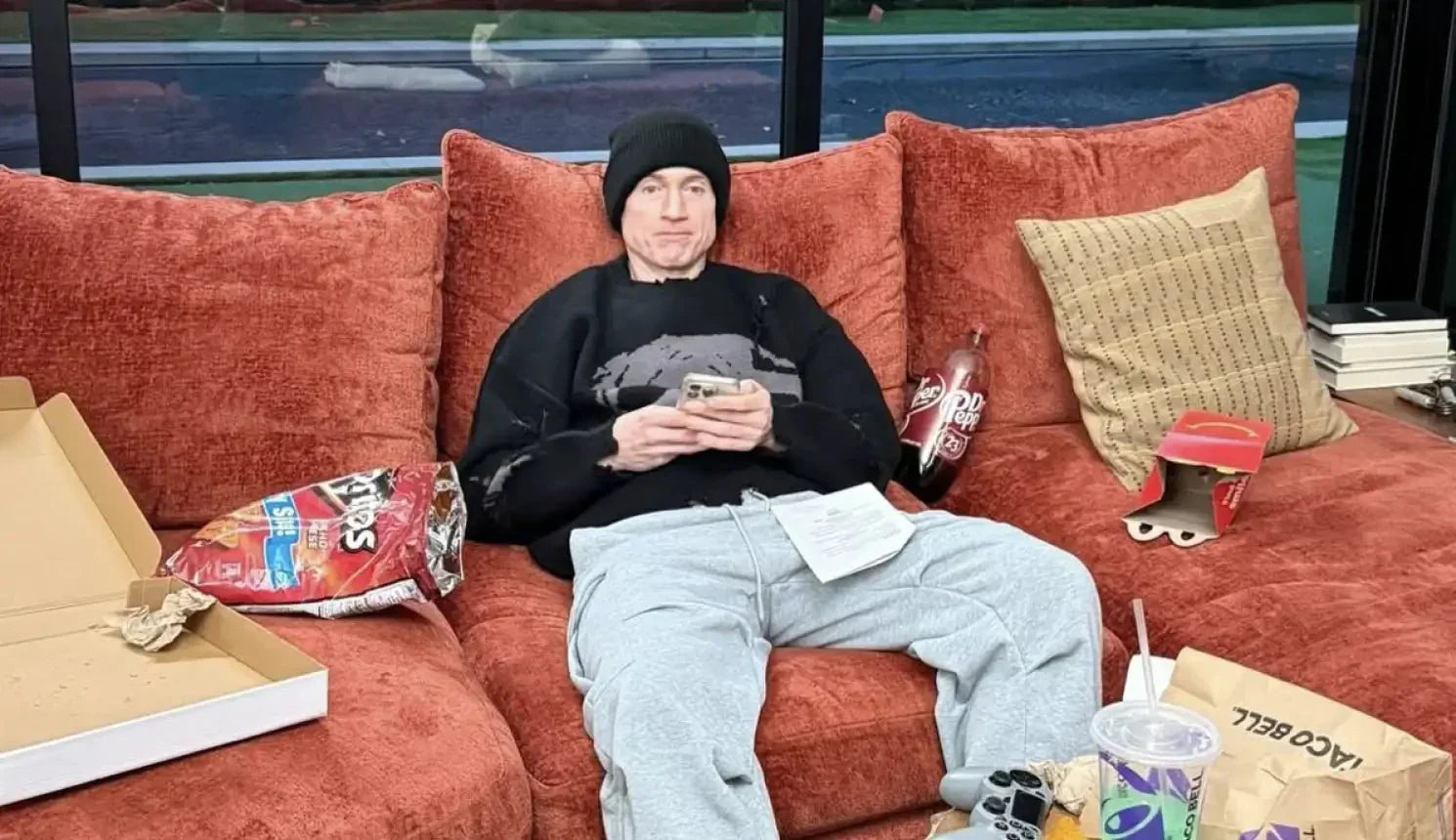 Bryan Johnson sitting on a couch surrounded by fast food, chips, and soda while looking at his phone.