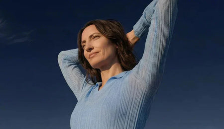 Woman in a blue shirt stretching her arms overhead against a clear blue sky.