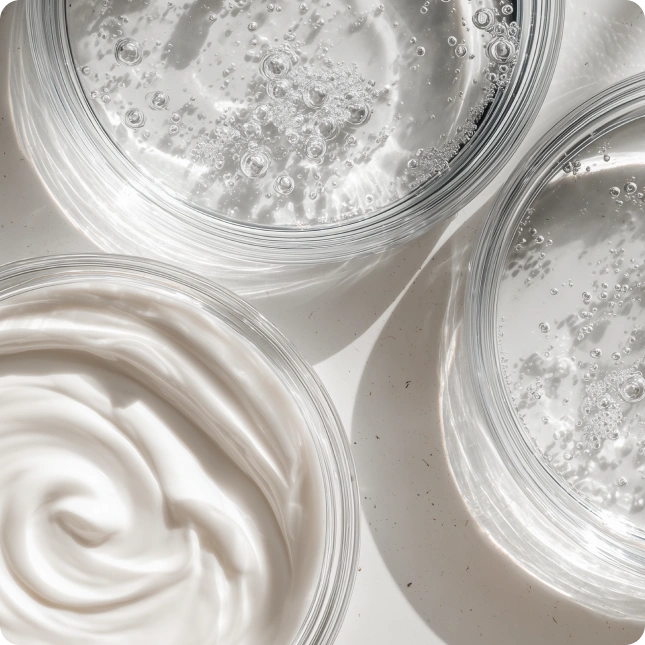 Close-up of three containers with white creamy skincare liquids and textures.