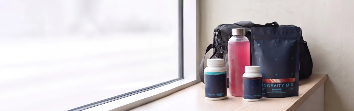 Blueprint Longevity Mix pouch with supplement bottles and a pink drink bottle beside a gym bag on a wooden windowsill