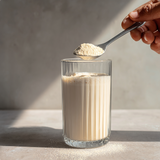 Glass of milk with a spoonful of powder being added against a neutral background