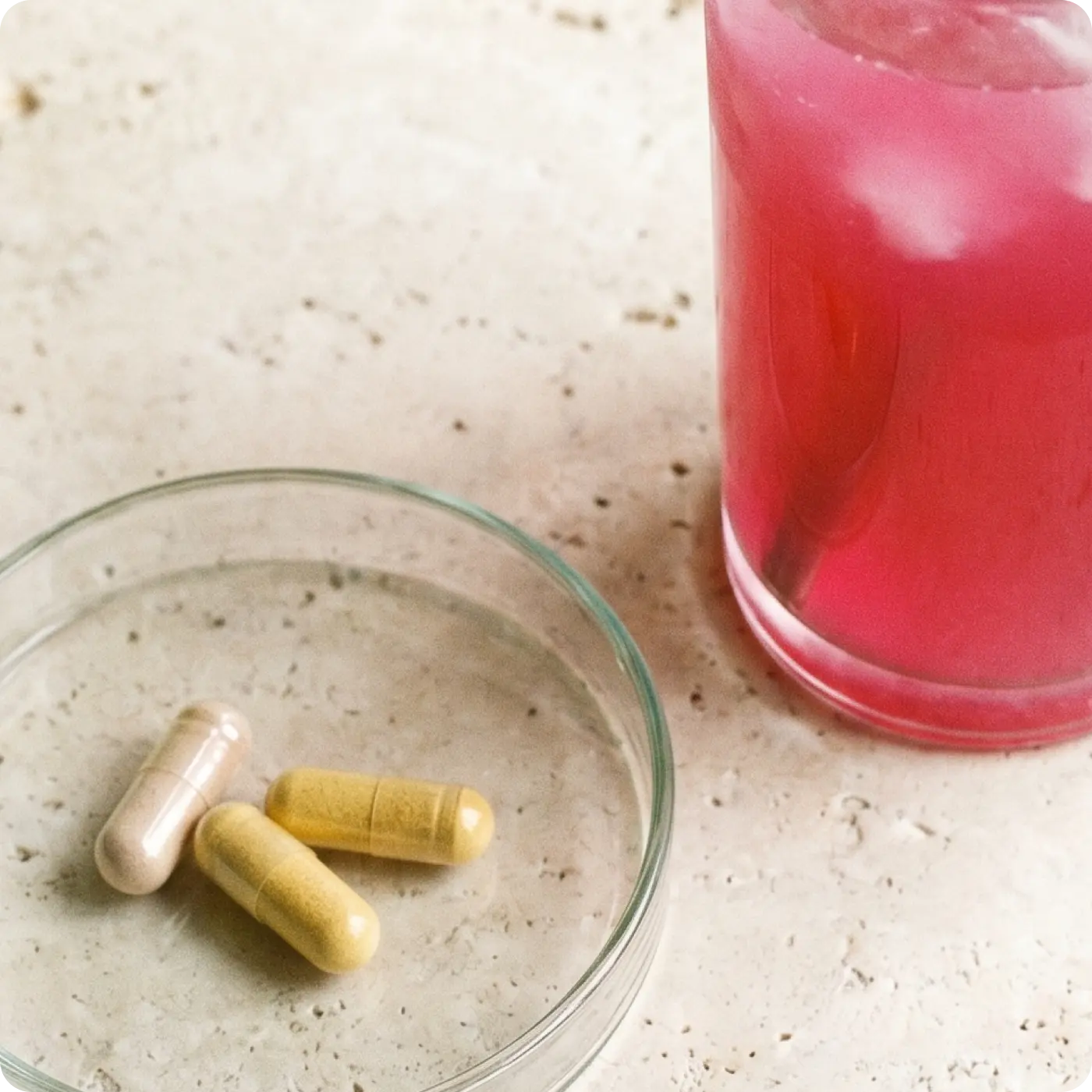 Two capsules on a glass plate next to a pink glass on a beige surface